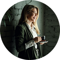 En person står inomhus vid en grå tegelvägg, håller en kopp kaffe och en fat. En grön växt syns till vänster i bilden.