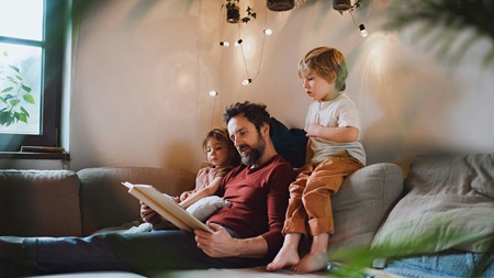 Vuxen sitter i en soffa och läser en bok med två barn i ett vardagsrum med varm belysning