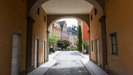 En vy genom en valvöppning som leder till en pittoresk gata med färgglada byggnader, träd och växter i ett lugnt bostadsområde.